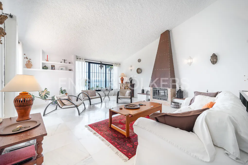 Bright living room with white walls, marble floors, and a tall copper fireplace. A white sofa, wood table, and chairs create a cozy seating area.
