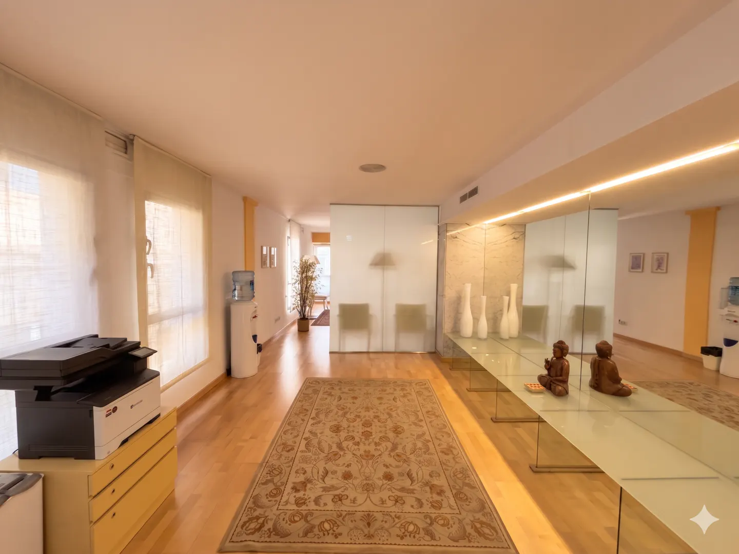 A bright office space with a long glass table, Buddha statues, and a patterned rug. A printer sits on a cabinet near the windows.