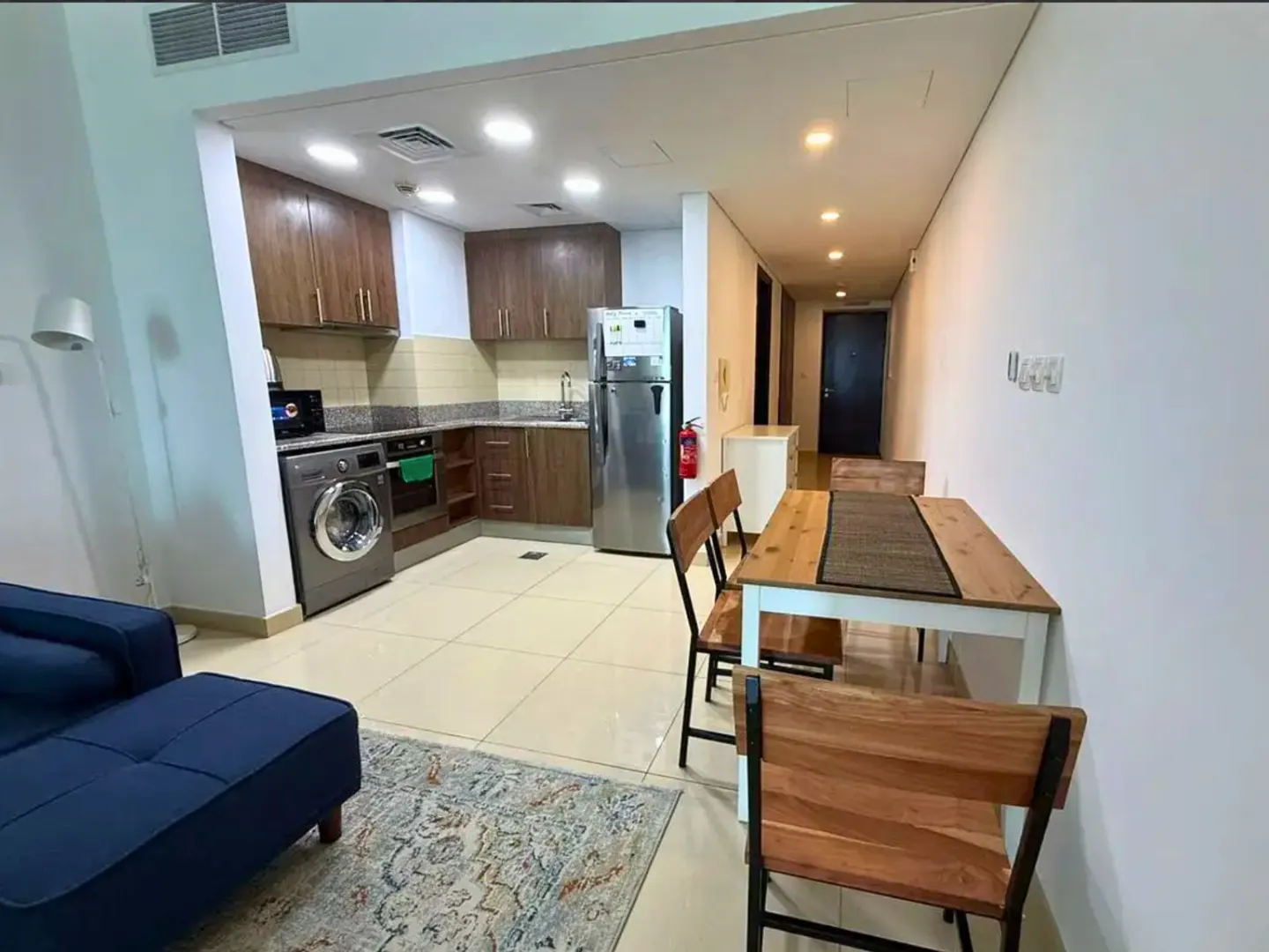A bright apartment interior with a blue sofa, patterned rug, dining table, and kitchen with stainless steel appliances.