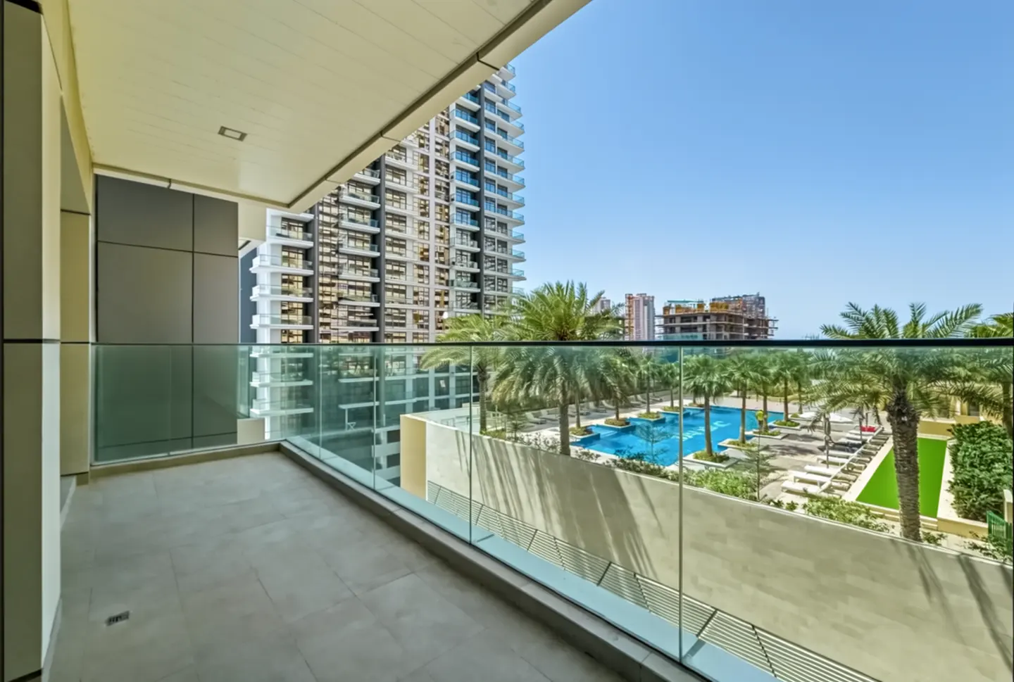 Balcony view of a pool and palm trees. High-rise building in the background under a blue sky. Glass railing.