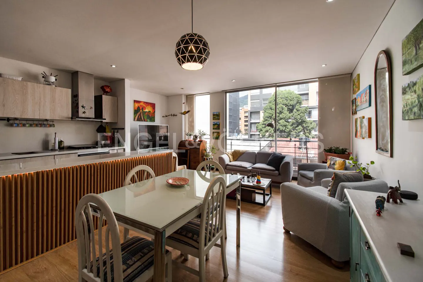 Open-concept living space with a dining table, kitchen, and seating area. Large windows offer city views. Hardwood floors and modern decor.