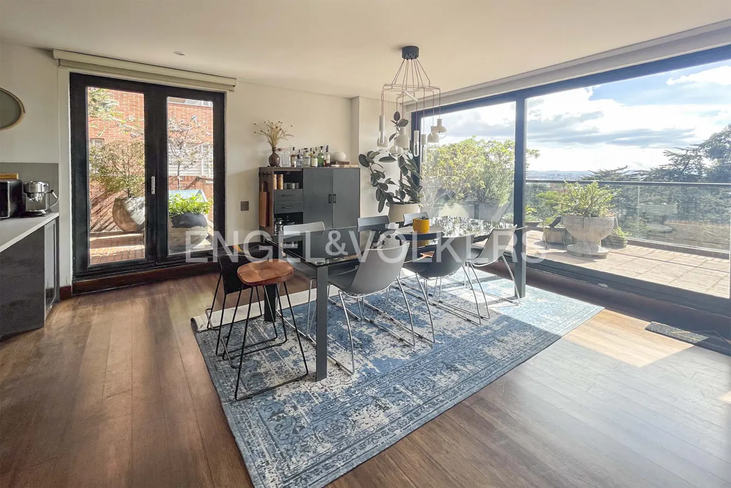 Bright dining area with wood floors, a blue rug, and a glass table with modern chairs. Sliding glass doors lead to a balcony with a view.