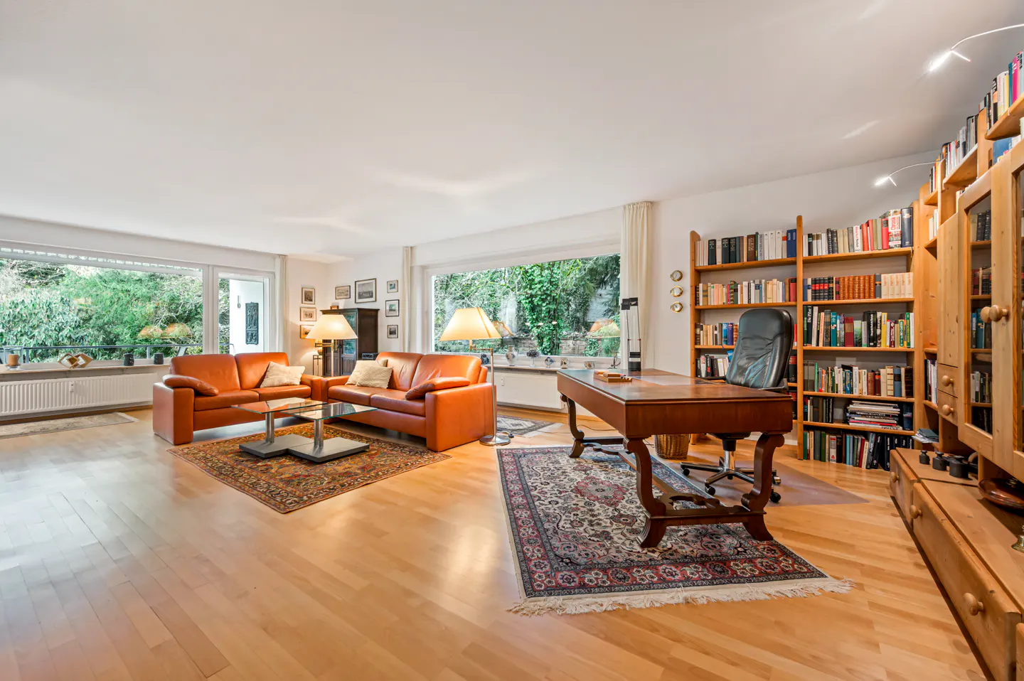 Bright living room with hardwood floors, orange leather sofas, a glass coffee table, and a wooden desk with bookshelves.