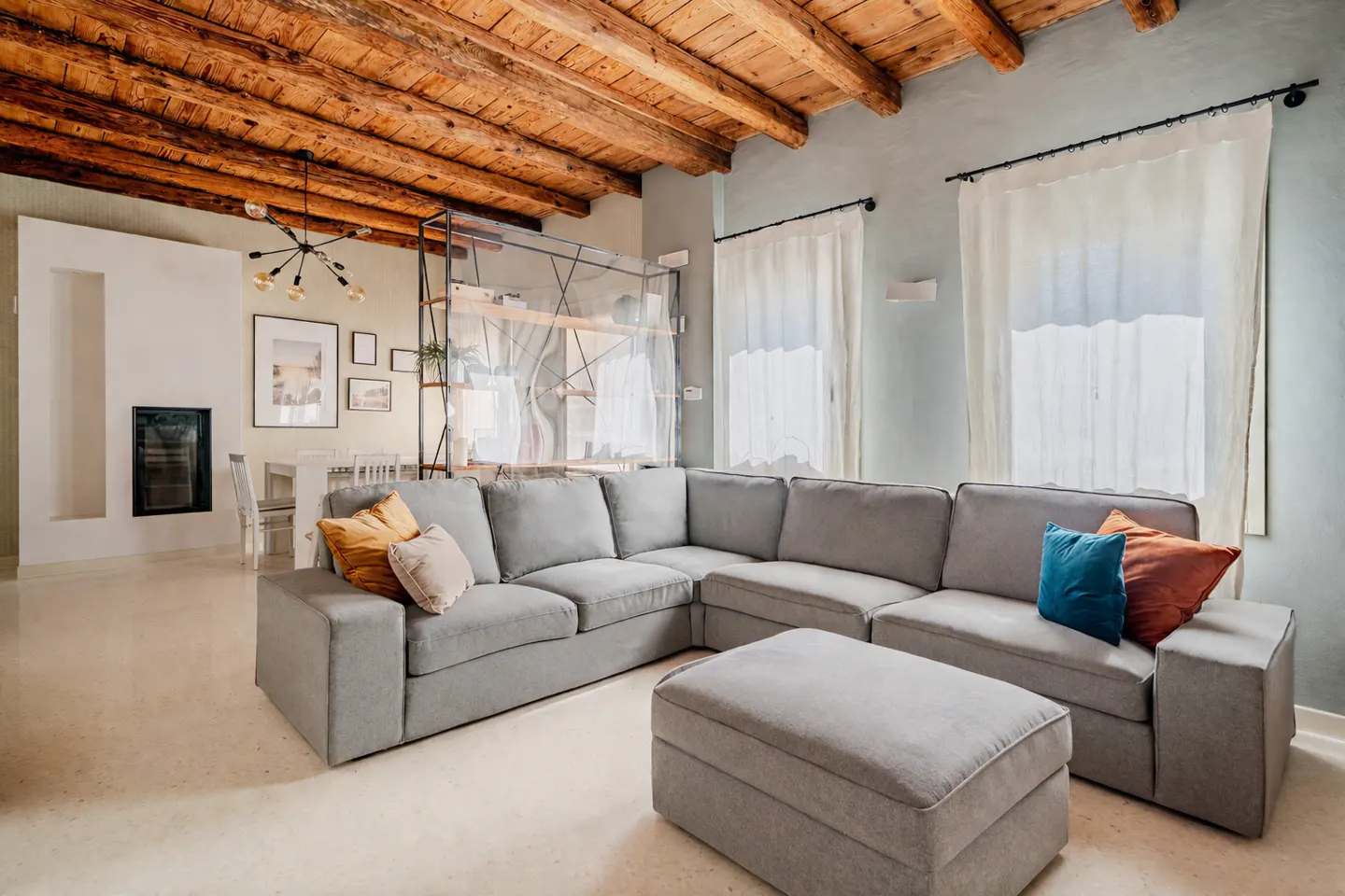 Living room with a gray sectional sofa, ottoman, and colorful pillows. Exposed wood beam ceiling and white curtains.