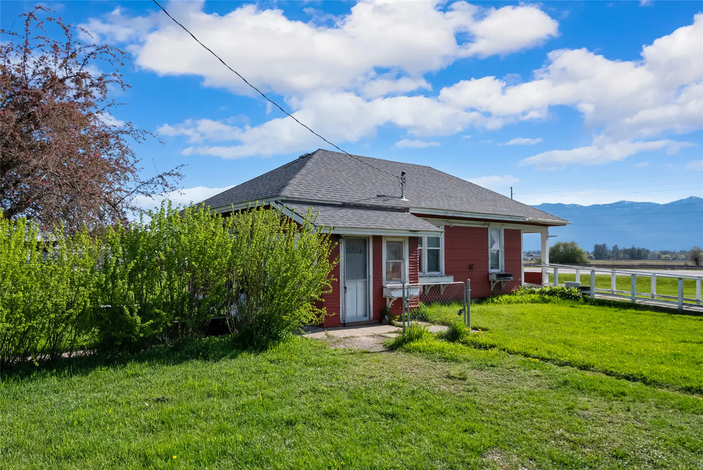 Red single-story house with a gray roof, green lawn, and blue sky with white clouds. Mountains are visible in the background.