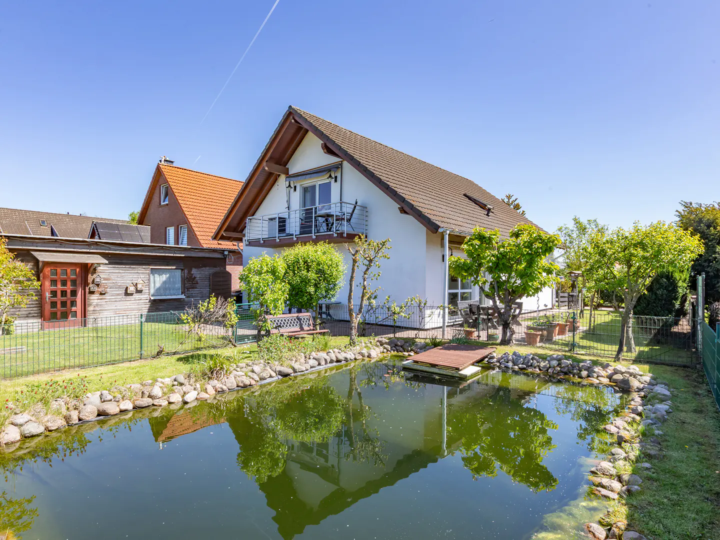 A white house with a brown roof overlooks a pond with a small wooden dock. Green trees and grass surround the pond.