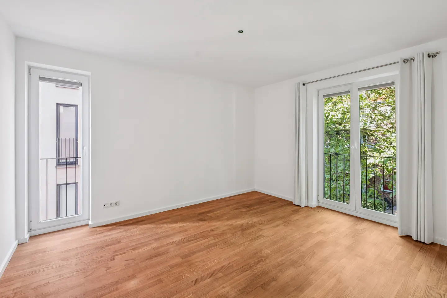 Empty room with white walls, hardwood floors, and two glass doors leading to balconies with views of trees and buildings.