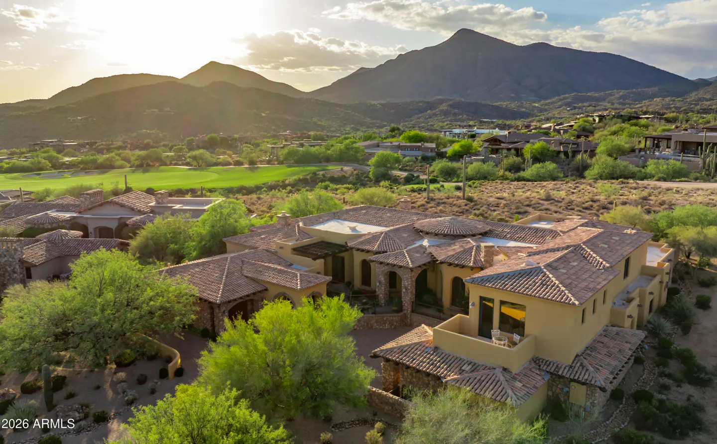 Aerial view of a tan, tile-roofed luxury home surrounded by green trees, with mountains and a golf course in the background.