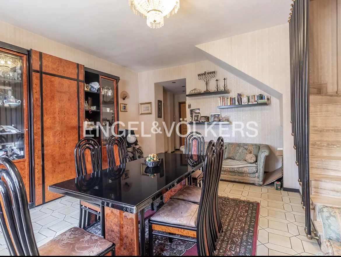 A dining room with a black table, wooden chairs, and a chandelier. A staircase and a sofa are in the background.