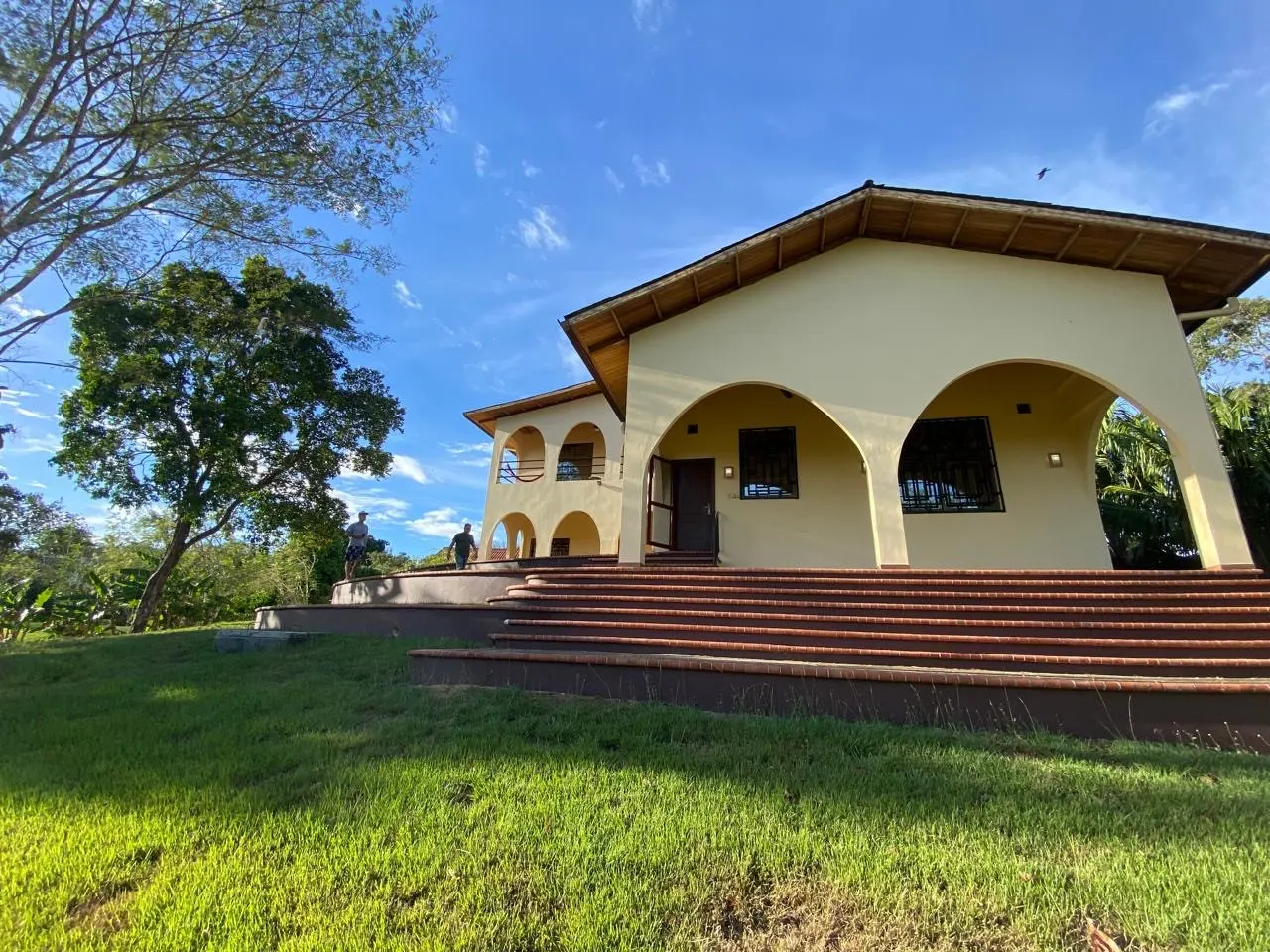 Beige house with arched porch and red brick steps on a green lawn under a blue sky. Two people are walking on the side.