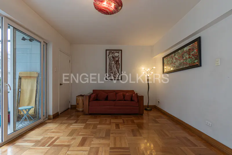 Living room with parquet floors, a red sofa, and a glass door to a patio. Artwork hangs on the white walls. A unique red lamp hangs from the ceiling.