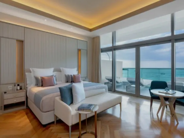 Bright bedroom with a king bed, white bench, and ocean view through large glass doors.
