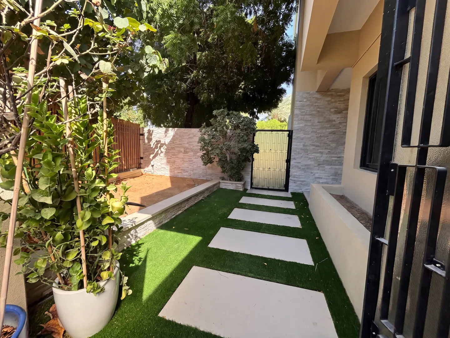 A side yard with a stone path on artificial grass, a potted plant, and a stone-tiled wall leading to a metal gate.