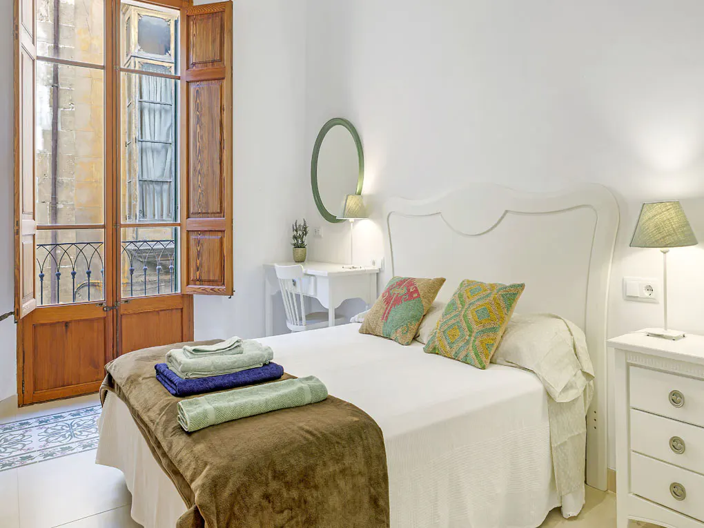 Bright bedroom with white walls, a bed with colorful pillows, and a desk near a window with wooden shutters.