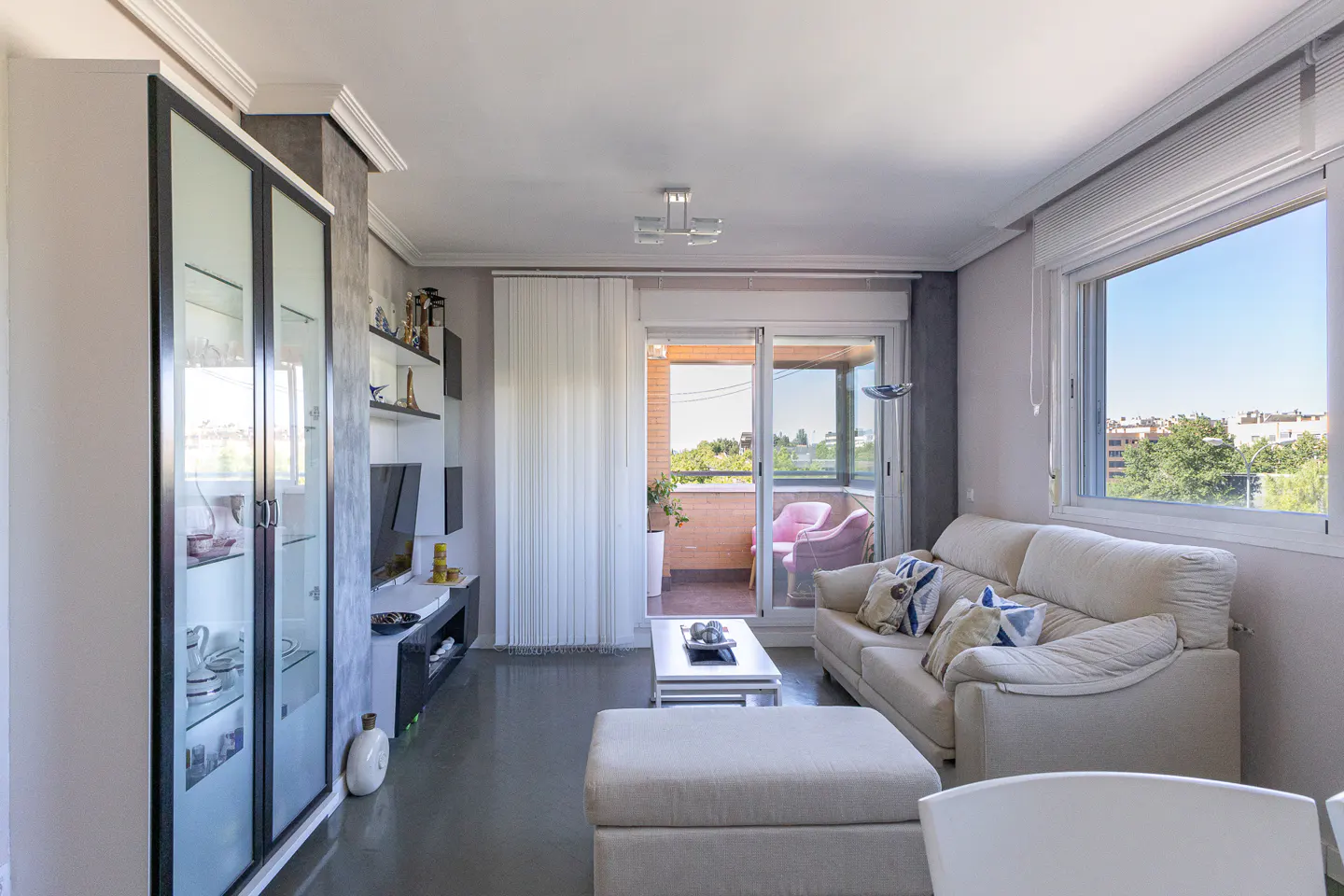 Bright living room with a beige sofa, matching ottoman, glass display cabinet, and a balcony with pink chairs.