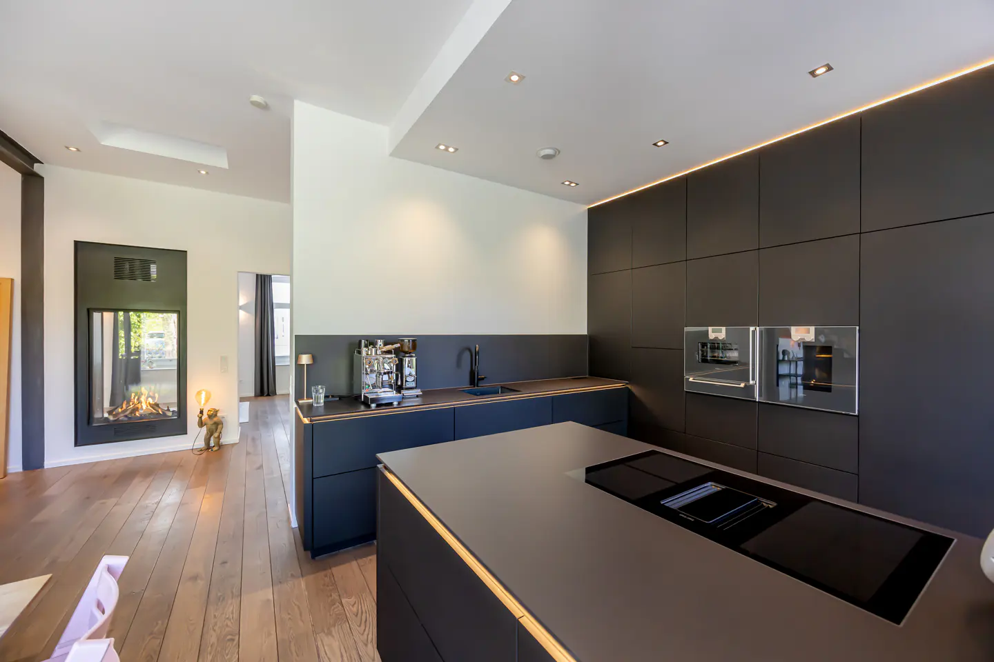 Modern kitchen with dark cabinets, wood floors, and a fireplace in the adjacent room. Stainless steel appliances and recessed lighting.