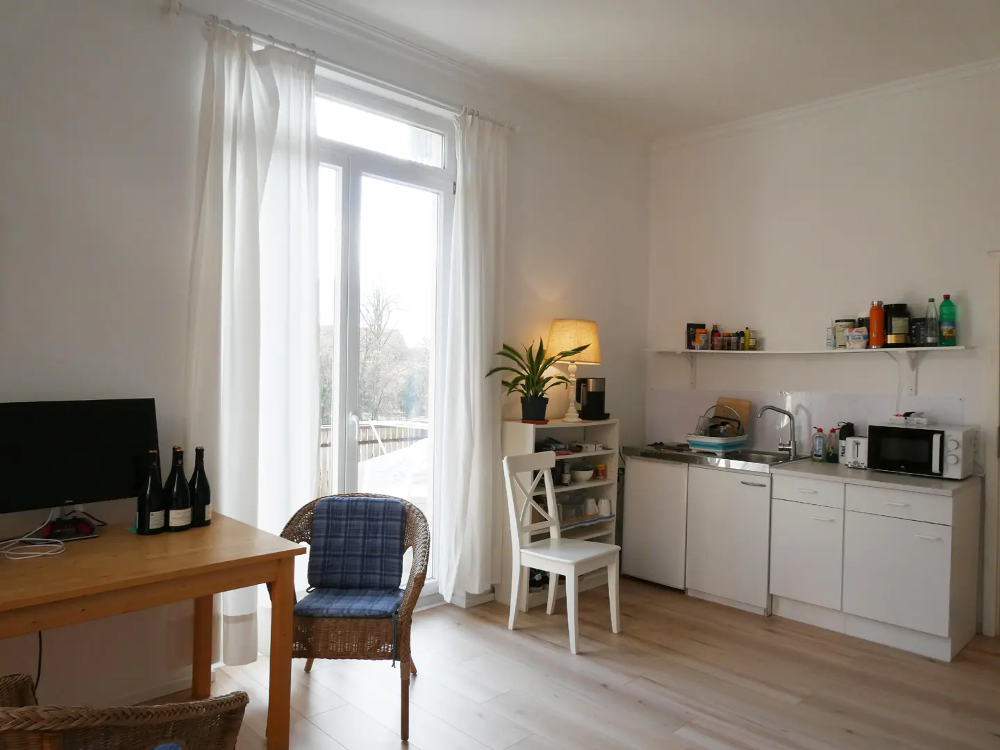 Bright studio apartment with white walls, wood floors, and a small kitchen. A table, chairs, and a large window with white curtains are visible.