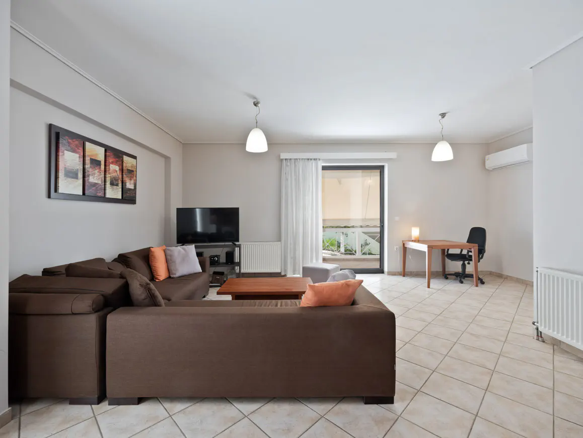 Living room with brown sectional sofa, TV, desk, and balcony with ocean view. Neutral colors and tile flooring.