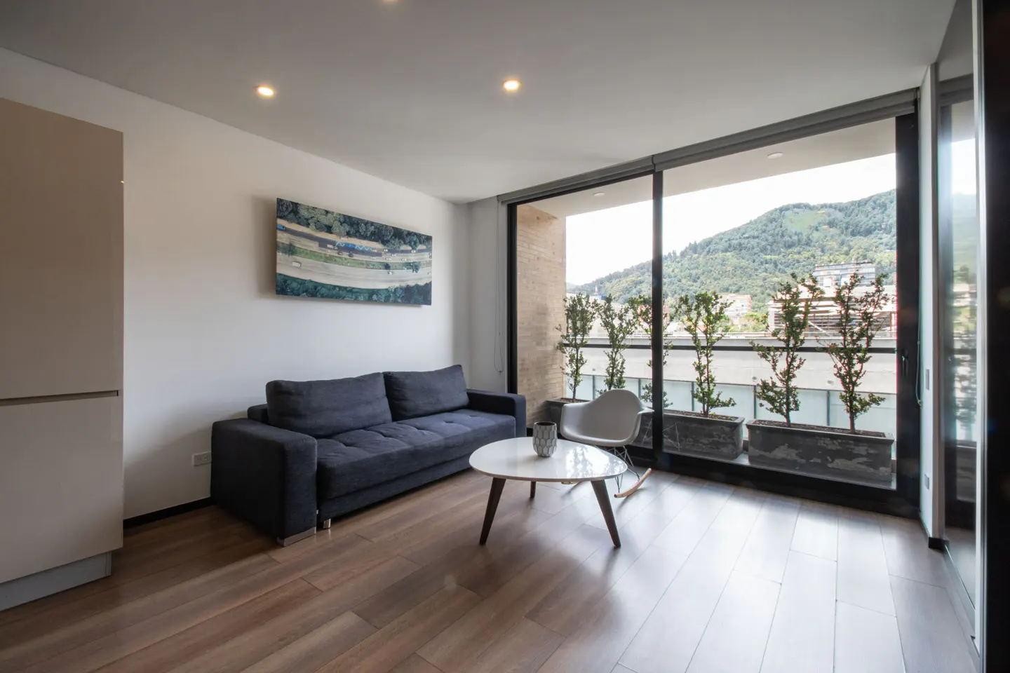 Bright living room with a gray sofa, white table, and balcony with mountain views. Artwork hangs on the wall.