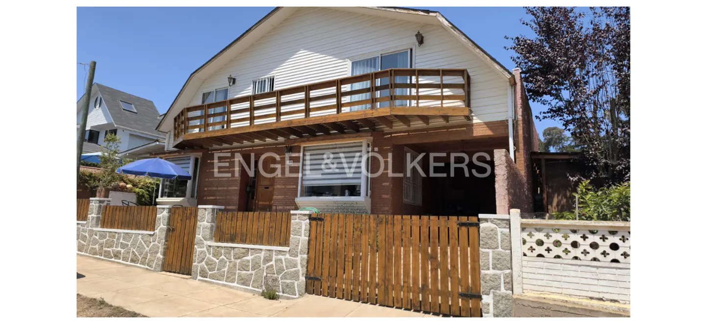 Two-story house with a wooden balcony, white siding, and a brick facade. A wooden fence with stone pillars surrounds the property. The Engel & Völkers logo is visible.
