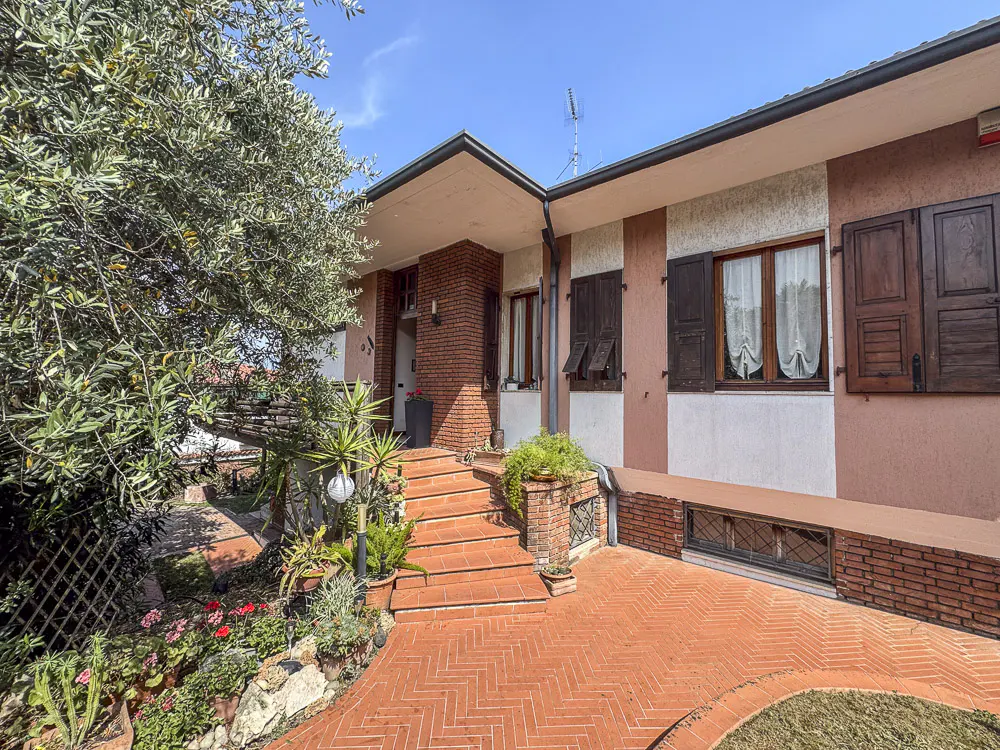 Exterior view of a two-story house with a brick walkway and garden. The house is painted in shades of pink and white, with brown shutters.