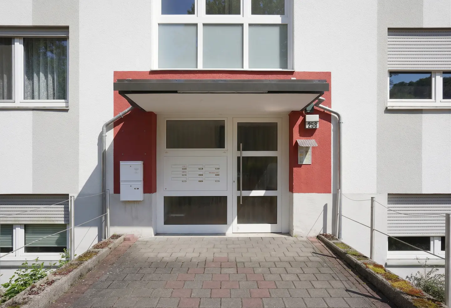 Apartment building entrance with white door, red accent wall, and brick ramp. Mailboxes and a small awning are visible.
