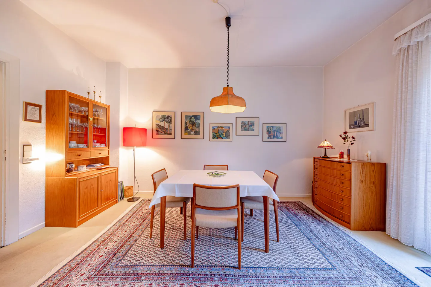 A dining room with a square table covered with a white tablecloth, surrounded by four chairs. A patterned rug covers the floor.