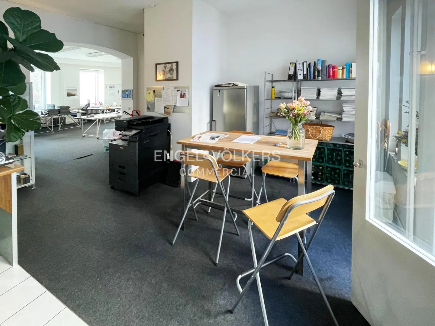 Office space with a wooden table, four chairs, and a black printer. Shelves with books and a stainless steel refrigerator are in the background.