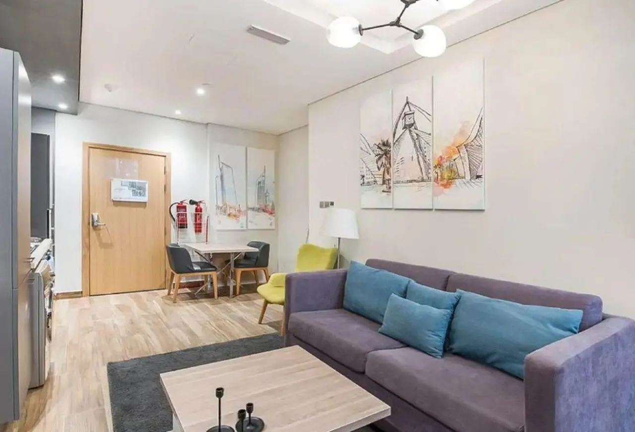 Living room with a purple sofa, blue pillows, a wooden coffee table, and a dining table with chairs. Artwork hangs on the wall.