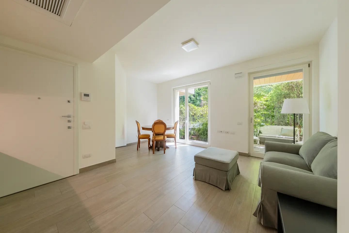 Bright living room with a gray sofa, ottoman, and wood dining set. Balcony doors open to a green outdoor space.