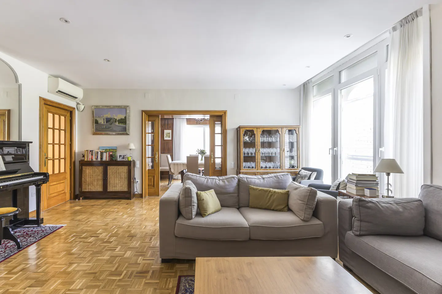 Bright living room with parquet floors, two grey sofas, a piano, and a view into a dining area through wooden doors.
