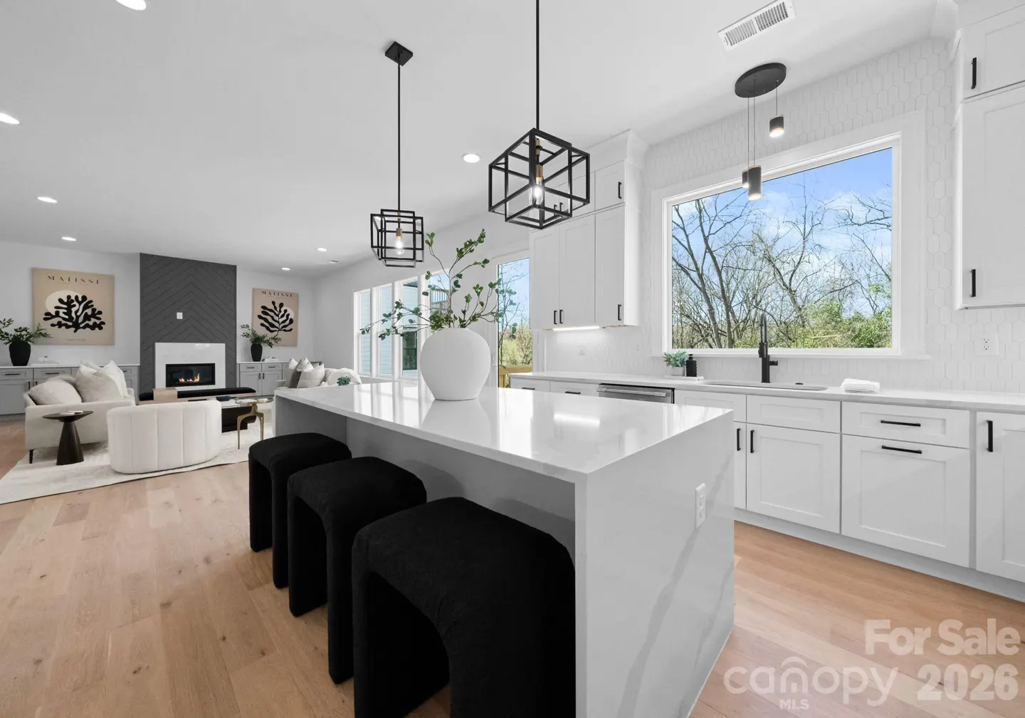 Modern white kitchen with a quartz island, black stools, and pendant lights, opening to a living room with a fireplace.