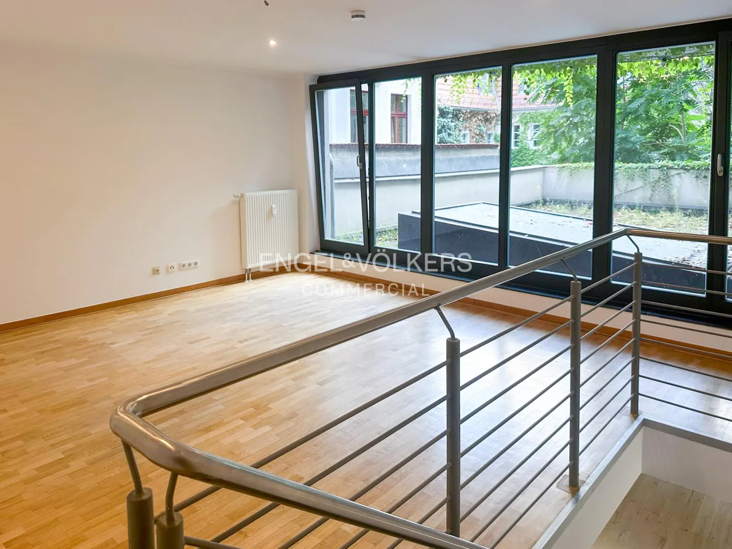 Bright room with wood floors, white walls, and large black-framed windows overlooking a green courtyard. A metal railing is in the foreground.