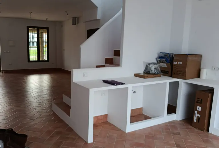 Unfurnished room with terracotta tile floor, white walls, and a staircase. A white built-in counter has boxes and a tablet on top. A window is visible in the background.