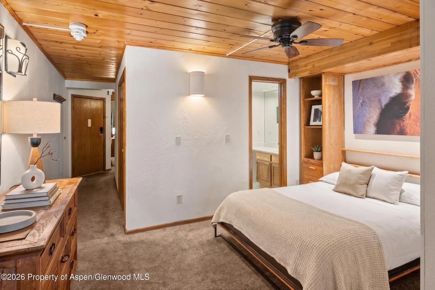Bedroom with wood ceiling, white walls, and brown carpet. A bed with white linens and a beige throw blanket is centered. A horse painting hangs above the bed.