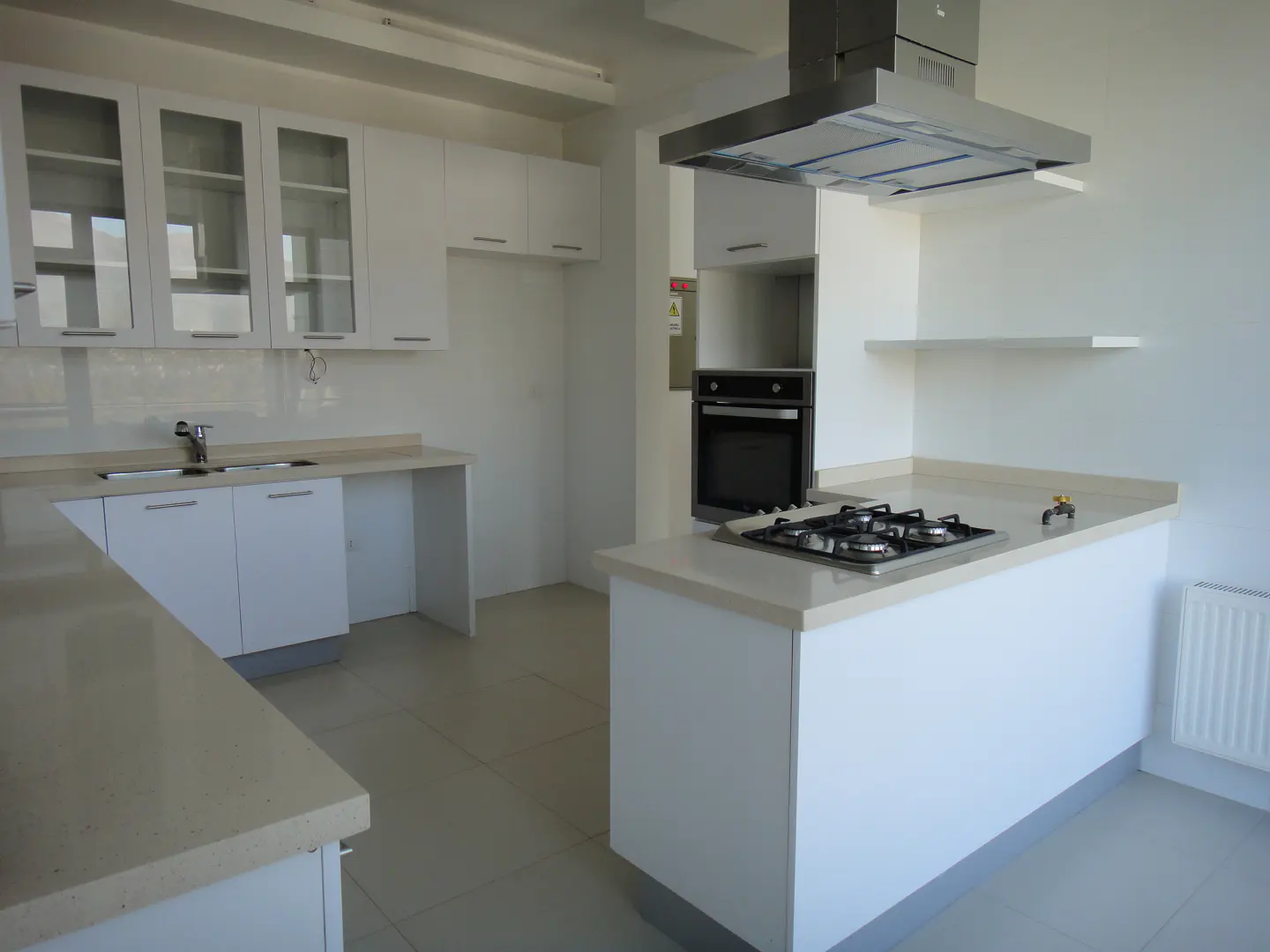 Bright, modern kitchen with white cabinets, countertops, and walls. A gas stovetop sits on the island, with a stainless steel range hood above.