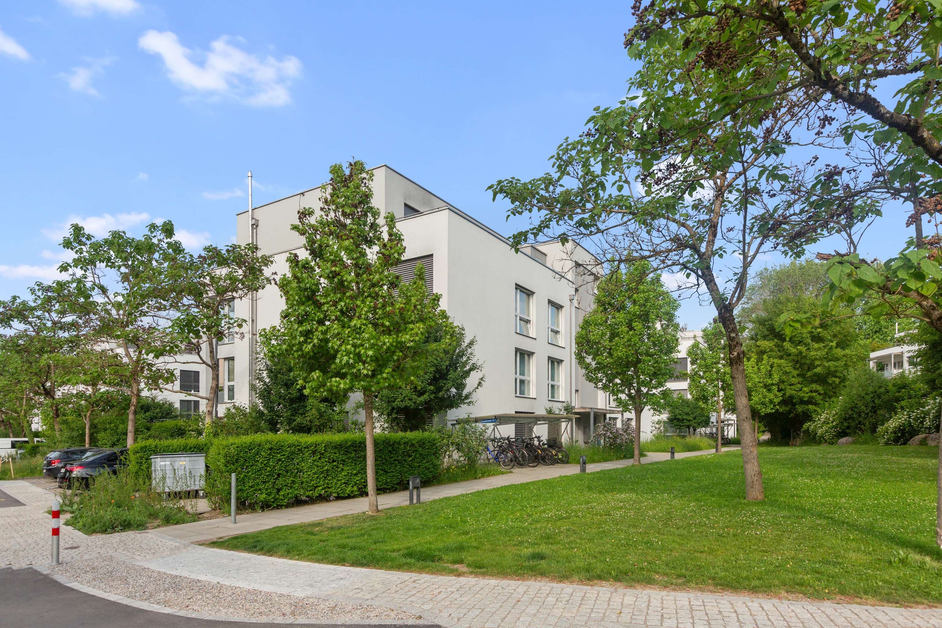 Modern white apartment building surrounded by green trees, a lawn, and a bicycle rack on a sunny day.