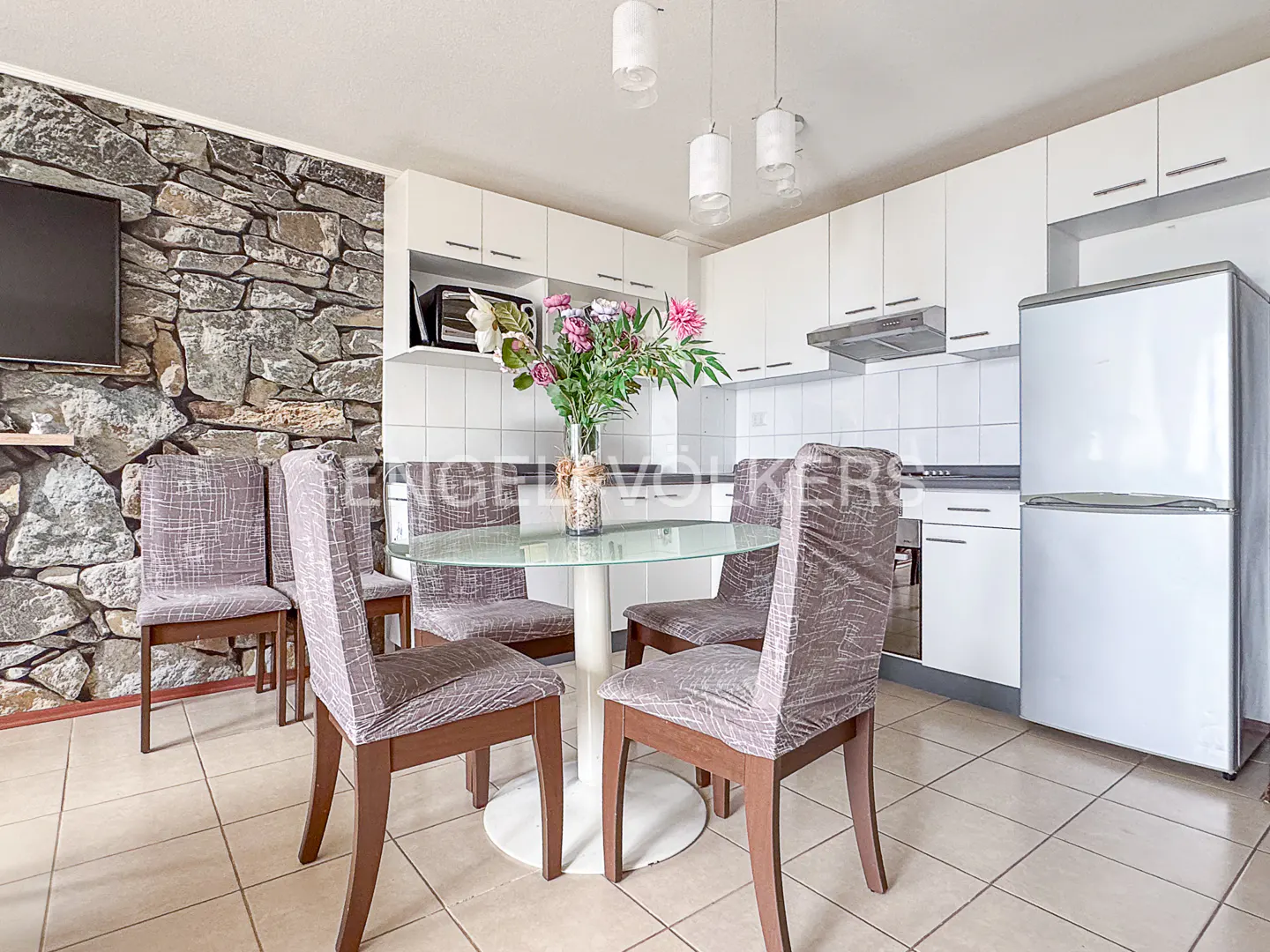 A kitchen with a stone wall, a TV, a glass table with flowers, and four chairs. The kitchen has white cabinets and a white refrigerator.