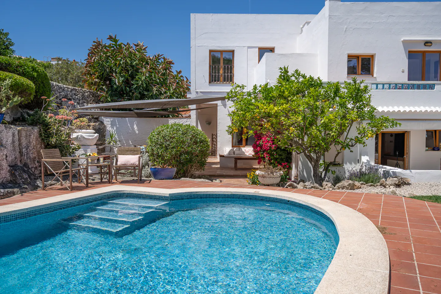 A white Mediterranean-style villa with a blue swimming pool, patio seating, and lush greenery under a clear blue sky.