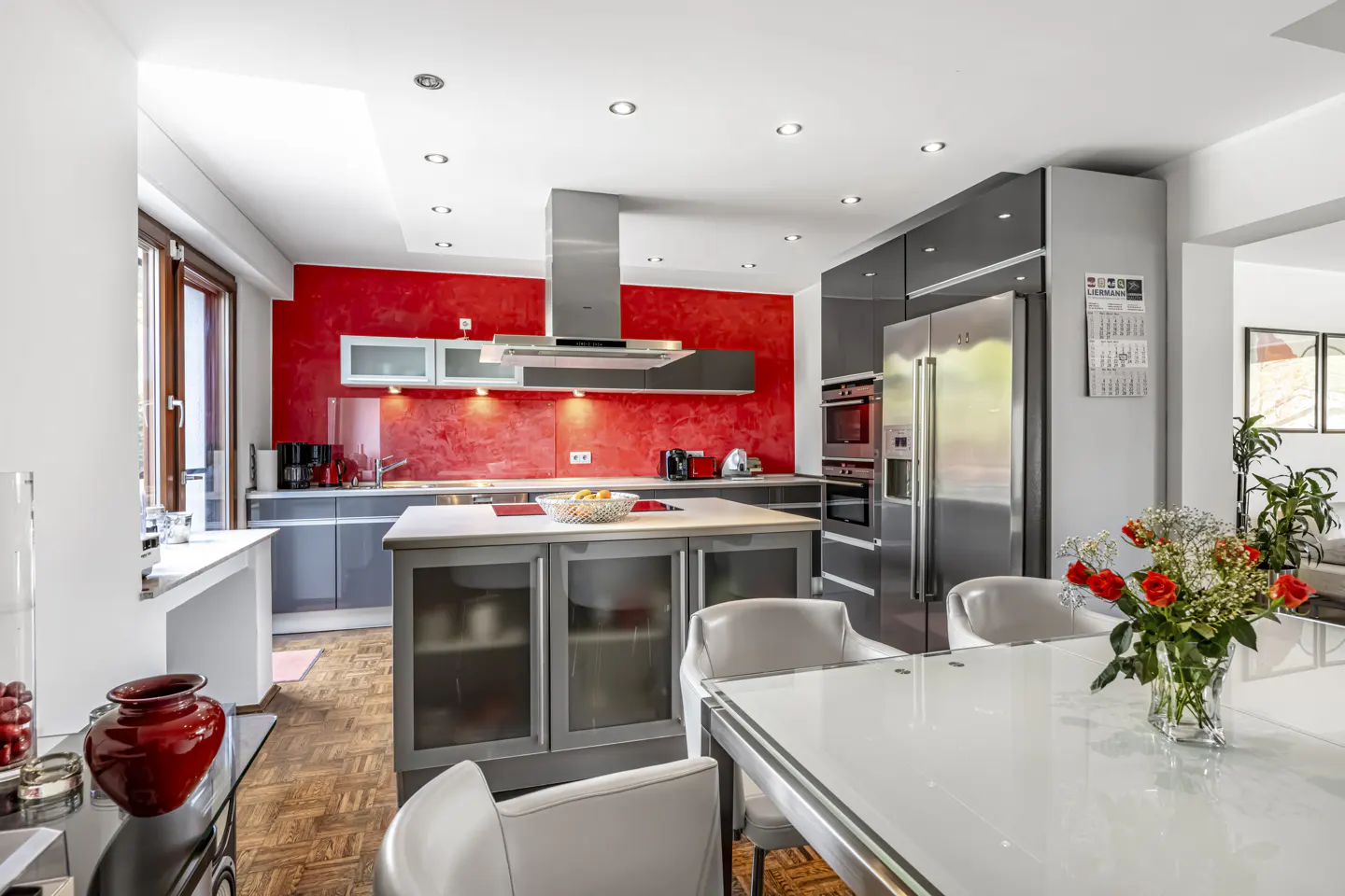 A modern kitchen with gray cabinets, stainless steel appliances, and a red accent wall. A white table with chairs sits in the foreground.