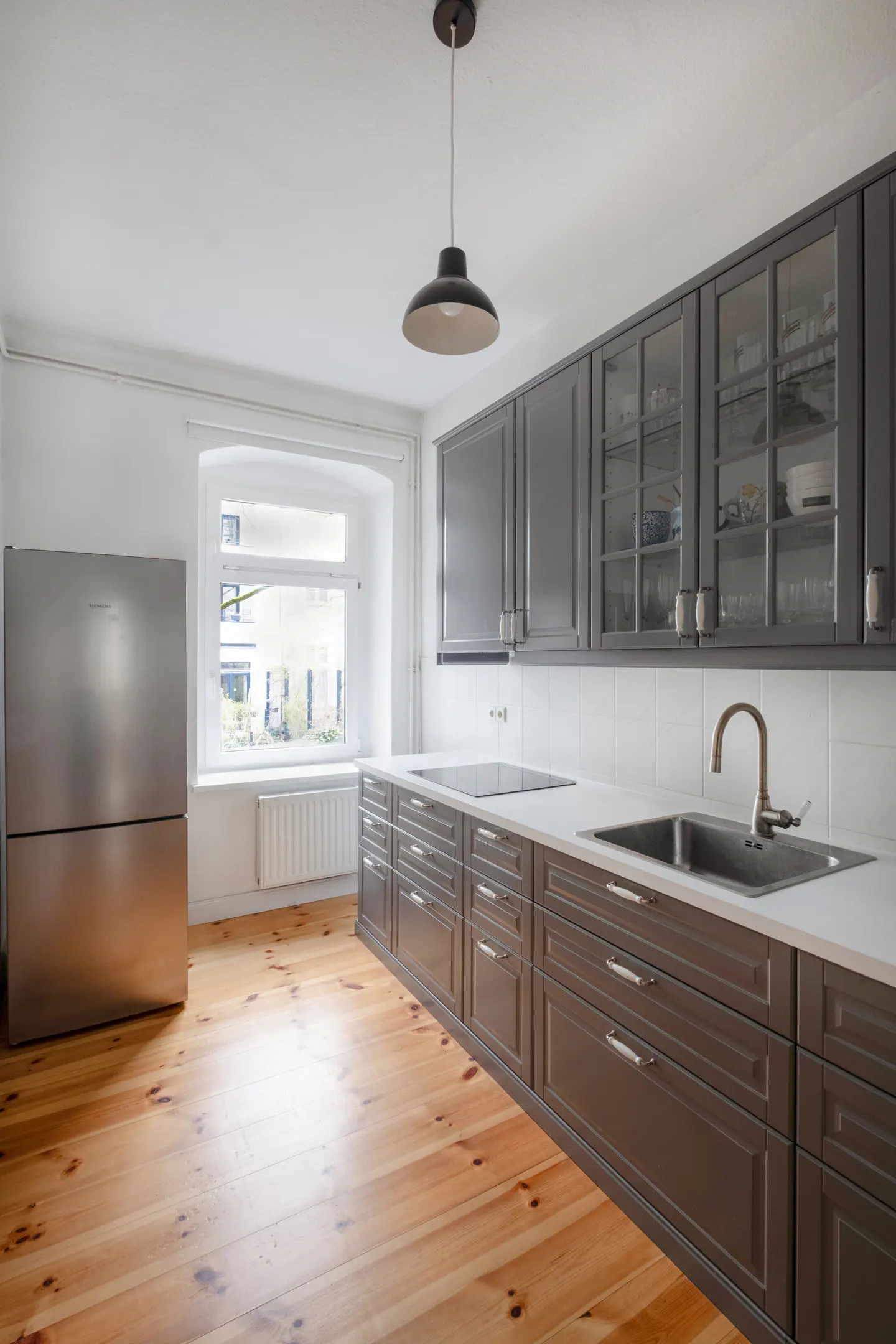 Bright kitchen with gray cabinets, stainless steel fridge, and light wood floors. A window overlooks a building.