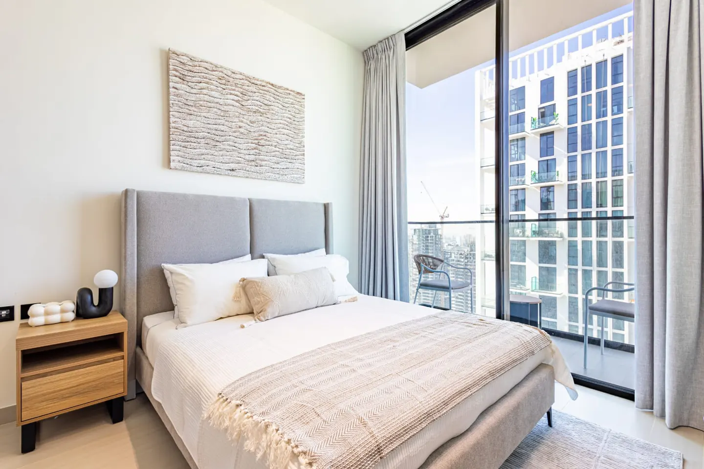 Bright bedroom with a gray headboard, white bedding, and a throw blanket. A balcony overlooks a city skyline.
