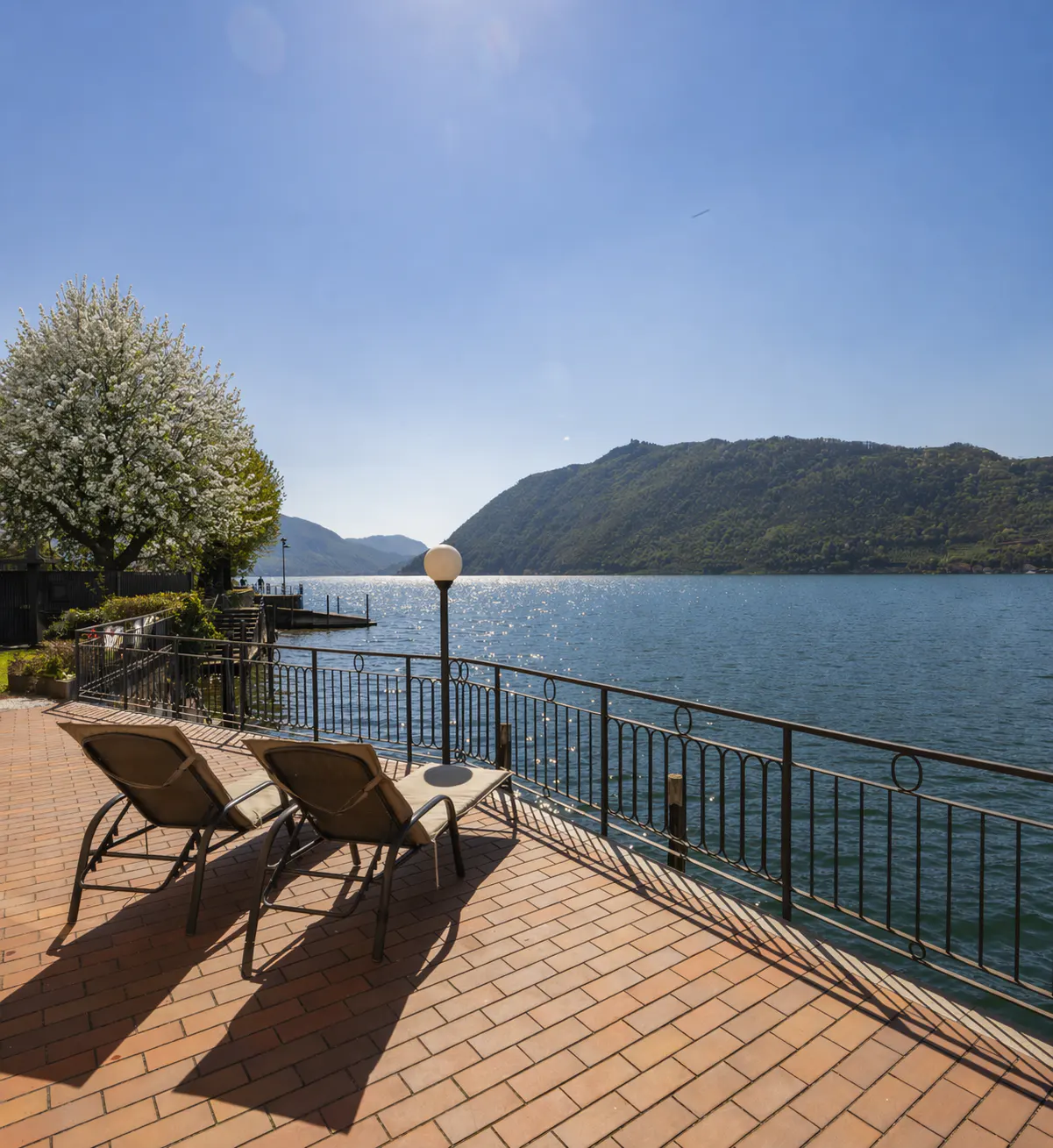 Two lounge chairs on a brick patio overlook a lake with mountains in the distance under a clear blue sky.
