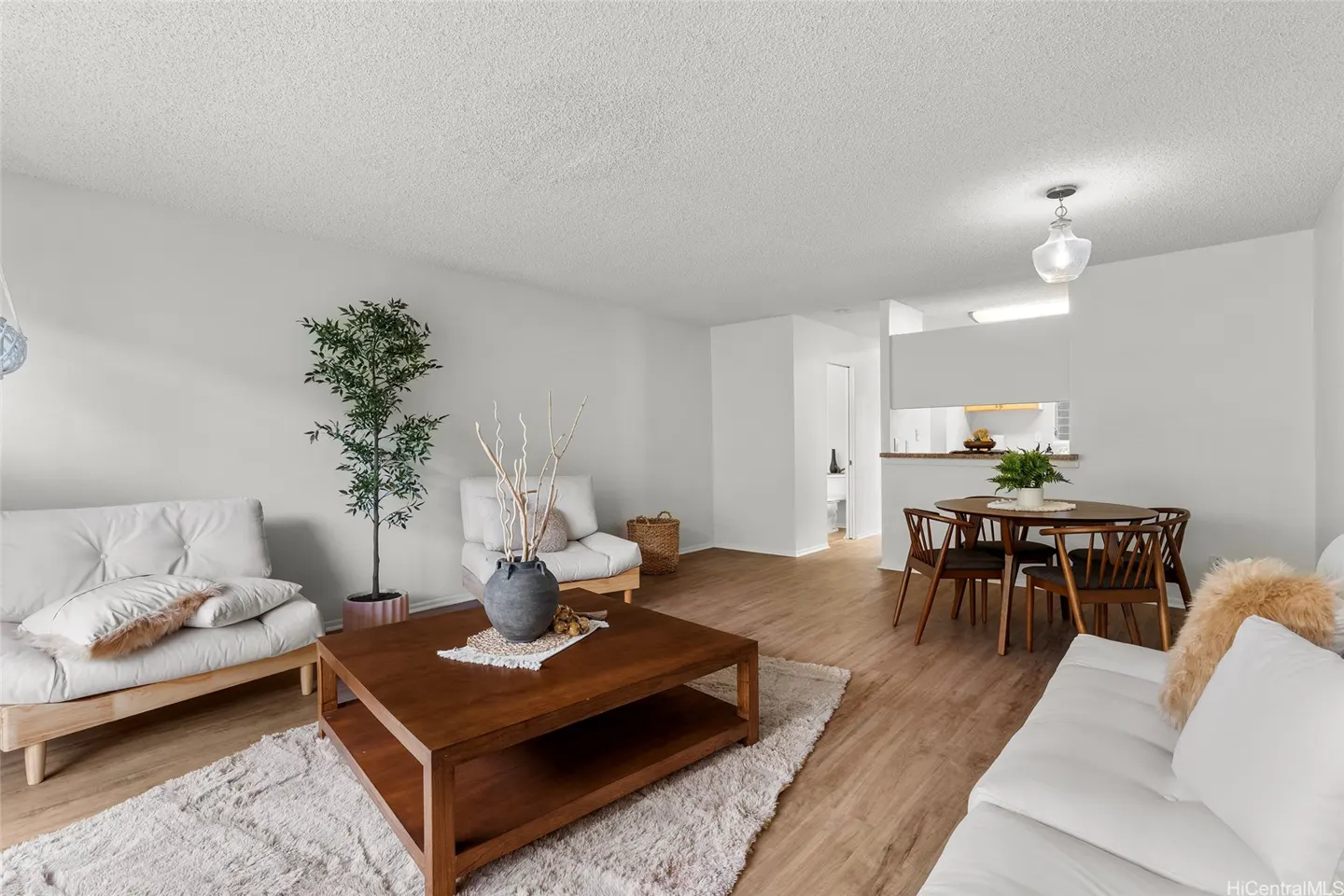 Bright living room with white sofas, a wooden coffee table, a tall potted plant, and a dining area with a wooden table.