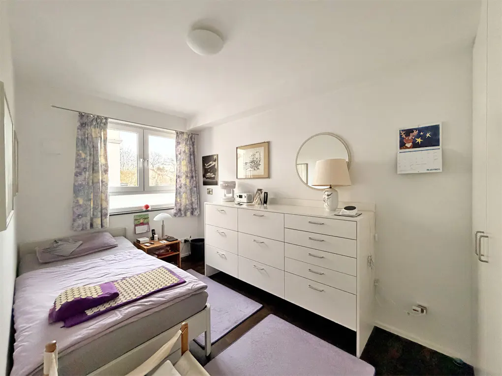 Bedroom with a single bed, white dresser, and lavender accents. Window with floral curtains lets in natural light.