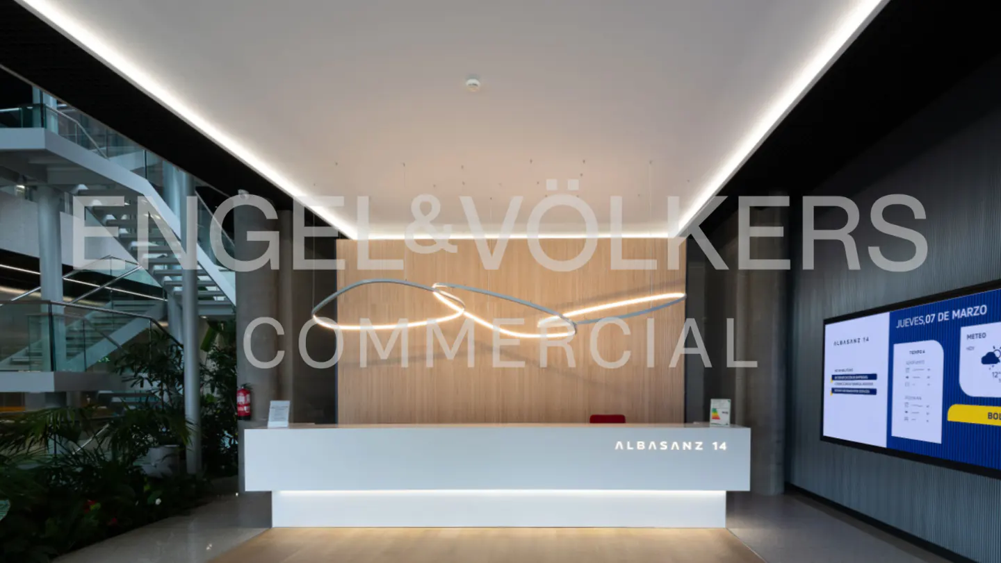 Engel & Völkers Commercial office lobby with a white reception desk, wood paneling, and a modern light fixture. A TV displays weather information.