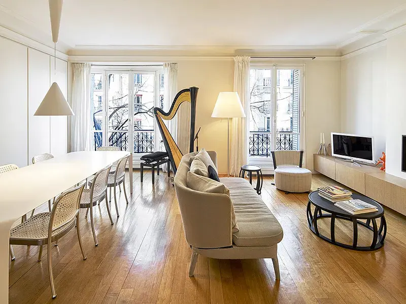 Bright living room with hardwood floors, a beige sofa, a harp, a dining area, and large windows with white curtains.