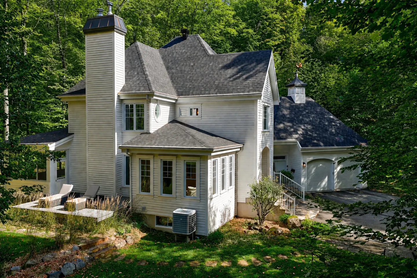 A white house with a gray roof is surrounded by green trees. The house has a tall chimney and a two-car garage.