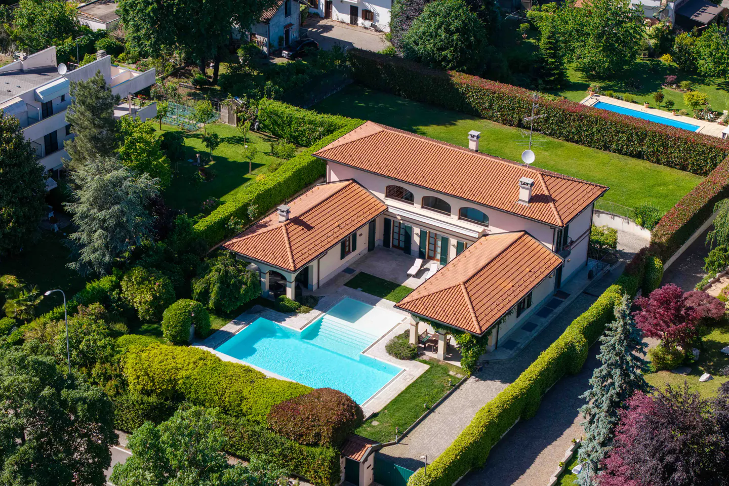 Aerial view of a pink house with a red tile roof, surrounded by green lawns, hedges, and a blue swimming pool.