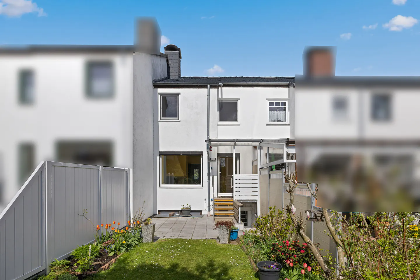 Backyard view of a white two-story house with a small lawn and flower beds under a blue sky.